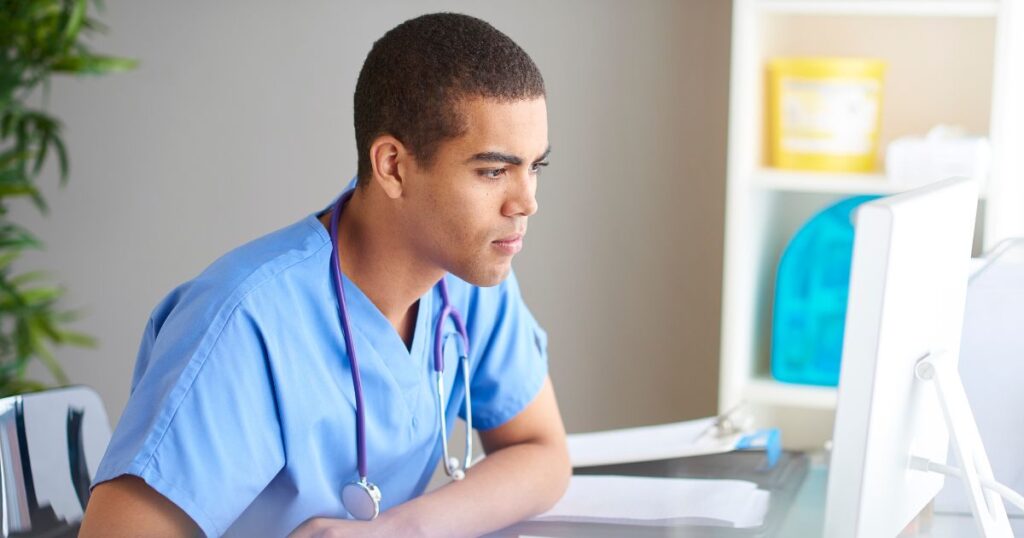 A young male nurse in blue scrubs is sitting and looking at a computer screen, learning about nursing license revocation.