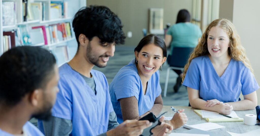 A group of nursing students wearing blue scrubs at a table discussing the causes of nursing license denial.